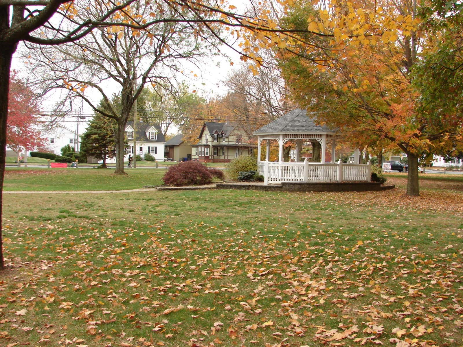 A white gazebo on a green with fallen leaves, under orange and yellow trees