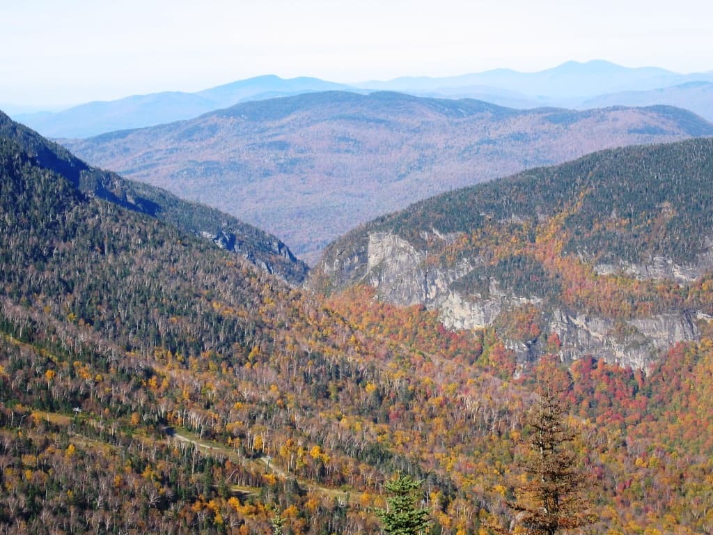 Steep forested slopes in fall colors dropping into a narrow mountain pass, with ridges beyond