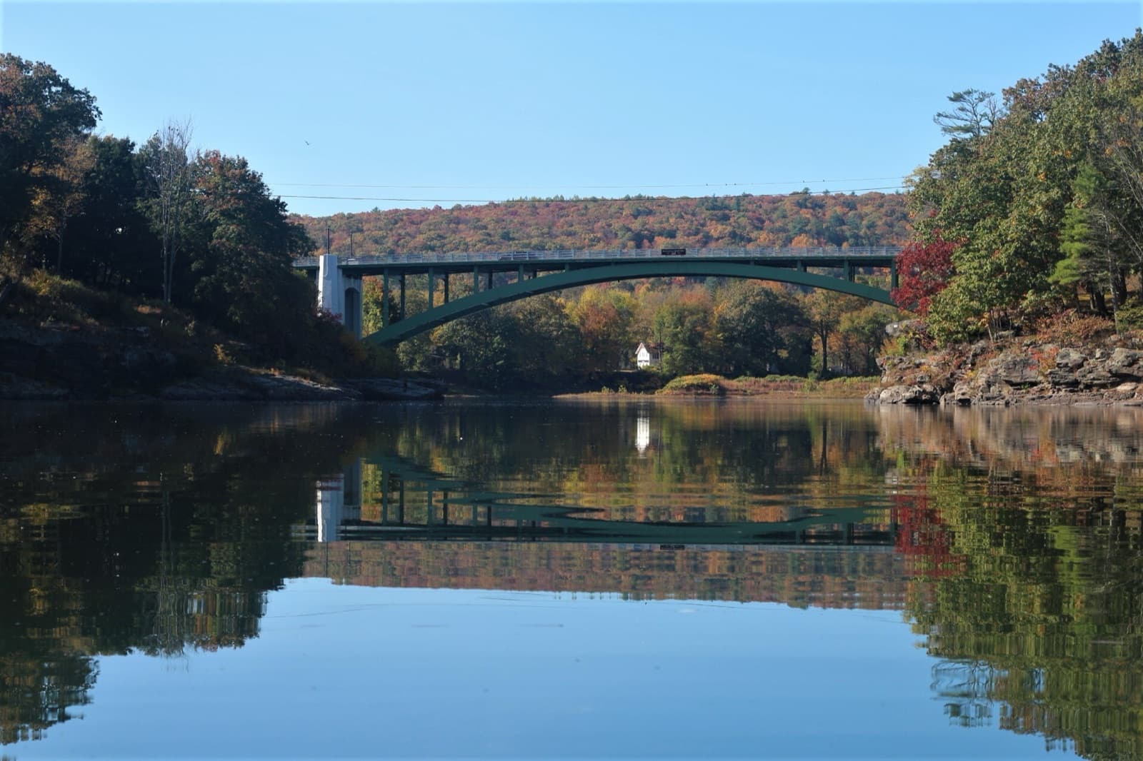A green arched bridge over a still river reflecting hills in fall color
