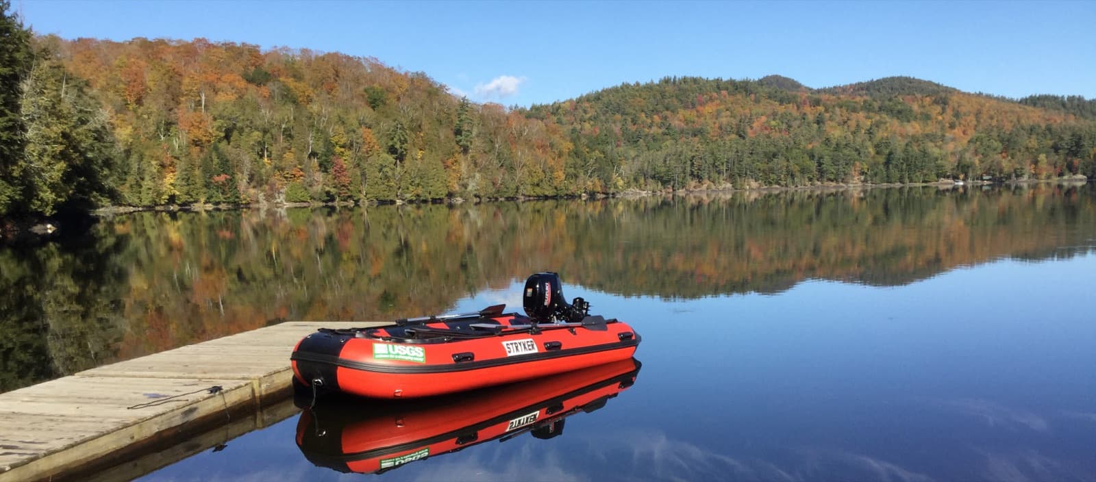 A lake reflecting hills in fall color, with a red USGS inflatable boat at a wooden dock
