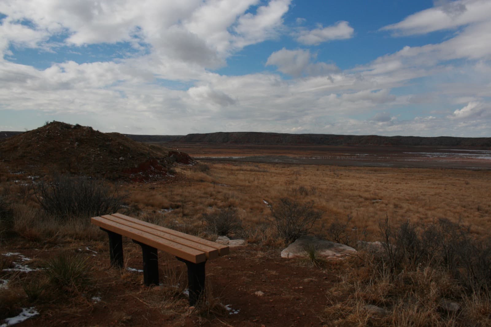 A wooden bench on a red-dirt rise above dry grassland, with the lake and flat-topped bluffs beyond under a cloudy sky