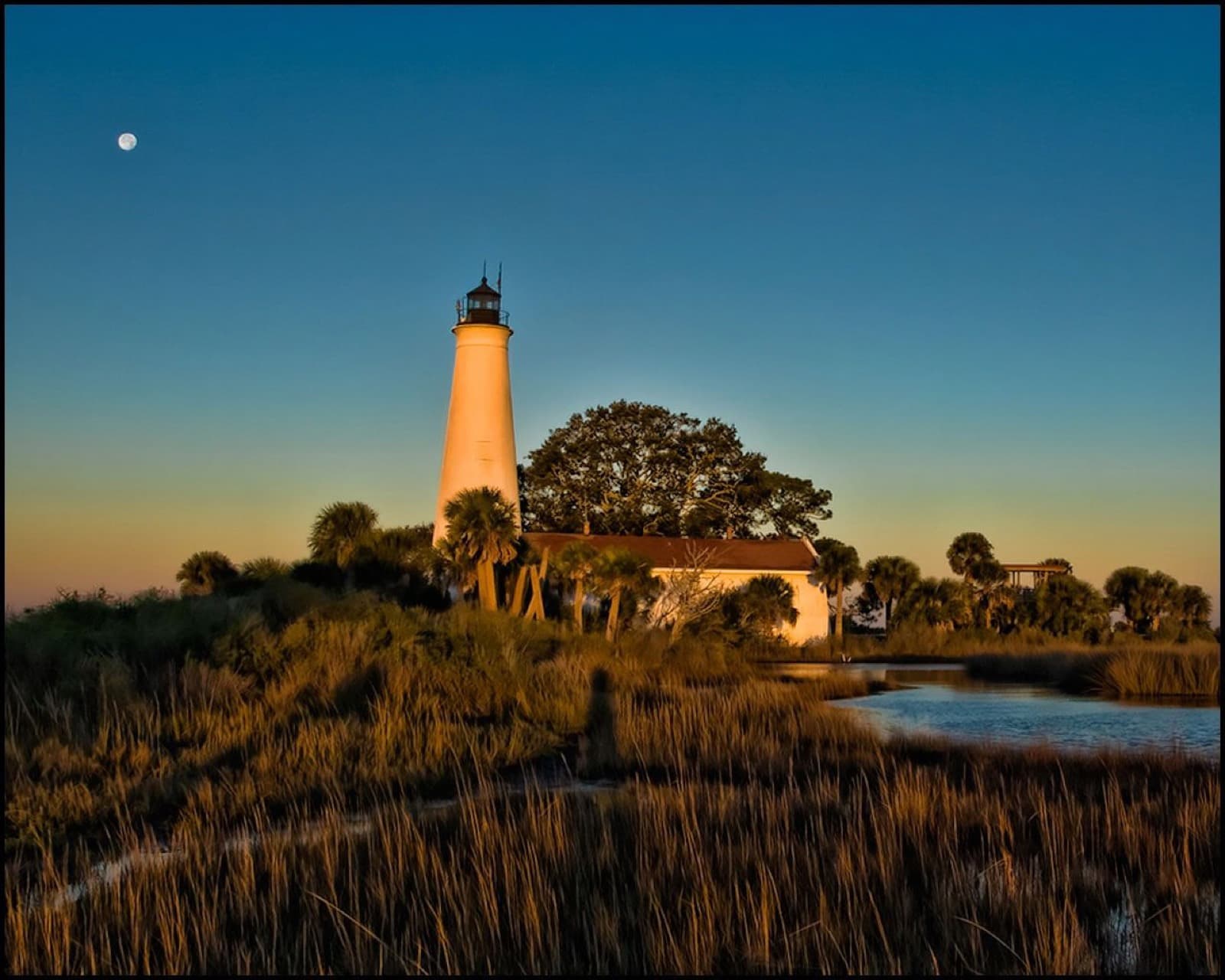 The white St. Marks Lighthouse and keeper's house in low golden light above marsh grass and a tidal creek, with the moon in a clear sky