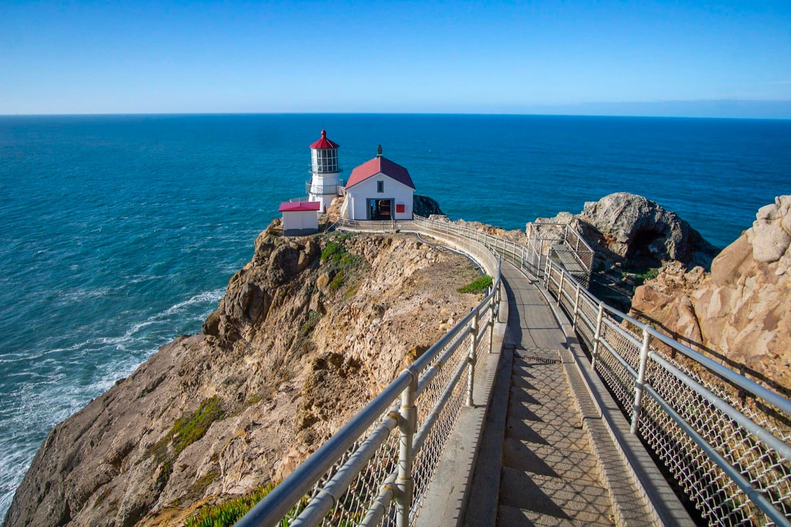 A long concrete staircase leads down to a white lighthouse with a red roof on cliffs above the ocean