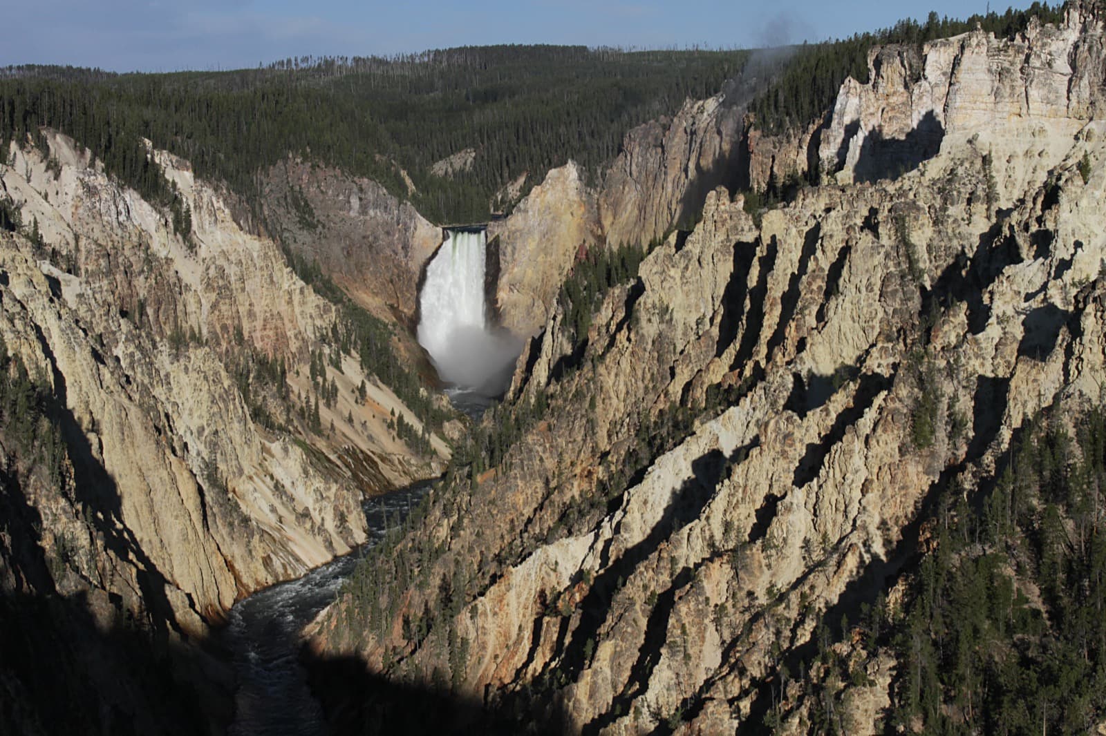 A tall waterfall at the head of a deep canyon with pale yellow rock walls, a river winding below and forest along both rims