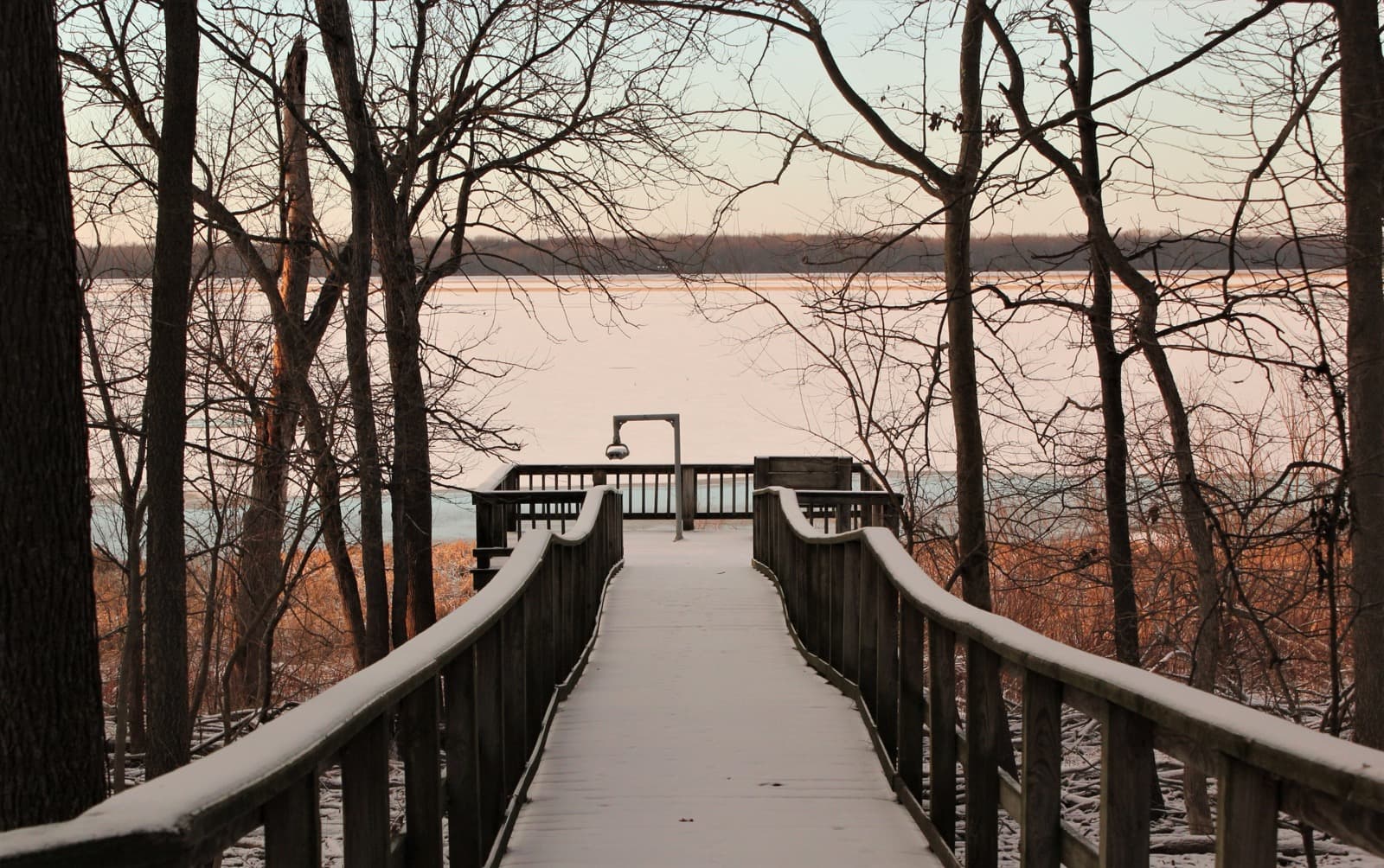 A snowy wooden boardwalk leads between bare trees to an overlook above a frozen lake at sunset