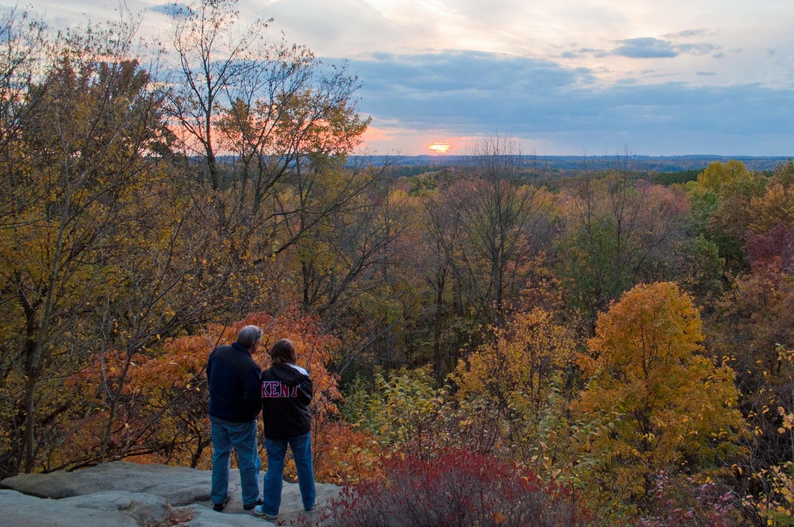 Two people stand at the edge of a gray rock cliff; in the distance, sunset and trees in fall colors
