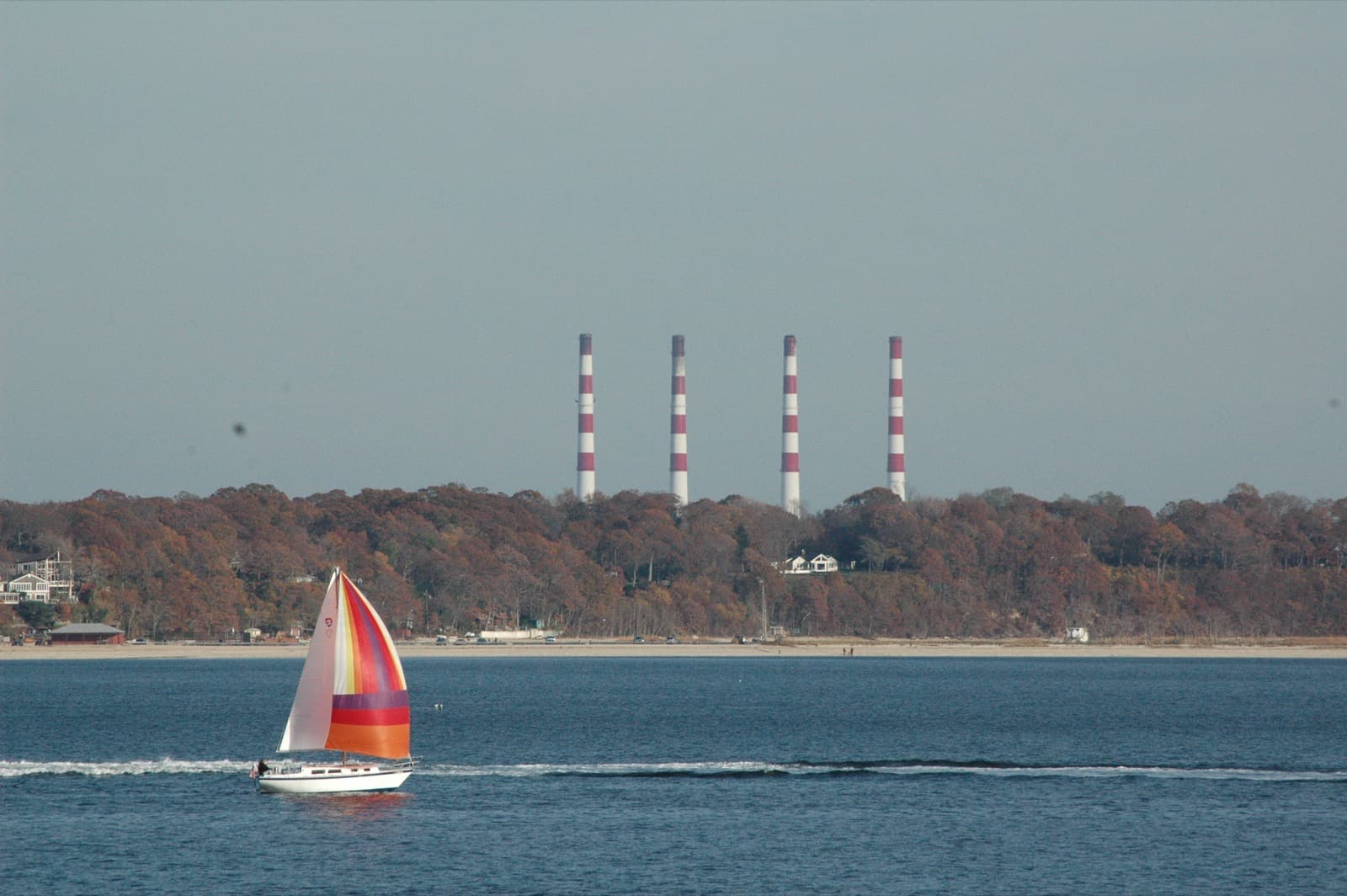 A sailboat with a bright striped sail on blue water, a wooded shoreline and four tall striped smokestacks in the distance
