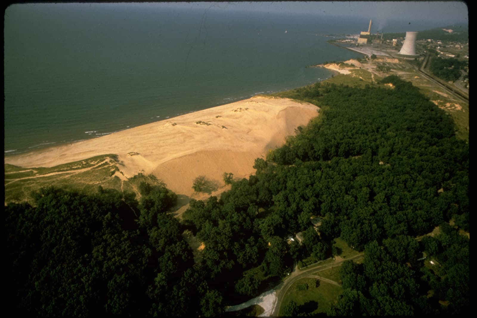 Aerial view of wooded dunes along a lake shore, with industrial buildings in the distance
