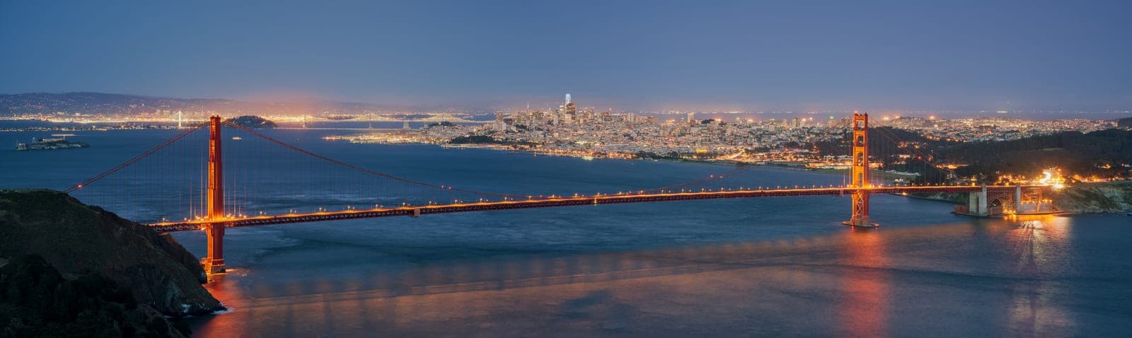 The lit Golden Gate Bridge crossing dark blue water toward the city lights of San Francisco at dusk