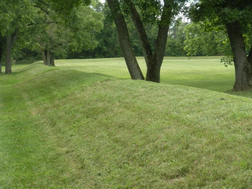 A long, low grass-covered earthen wall curving between large shade trees