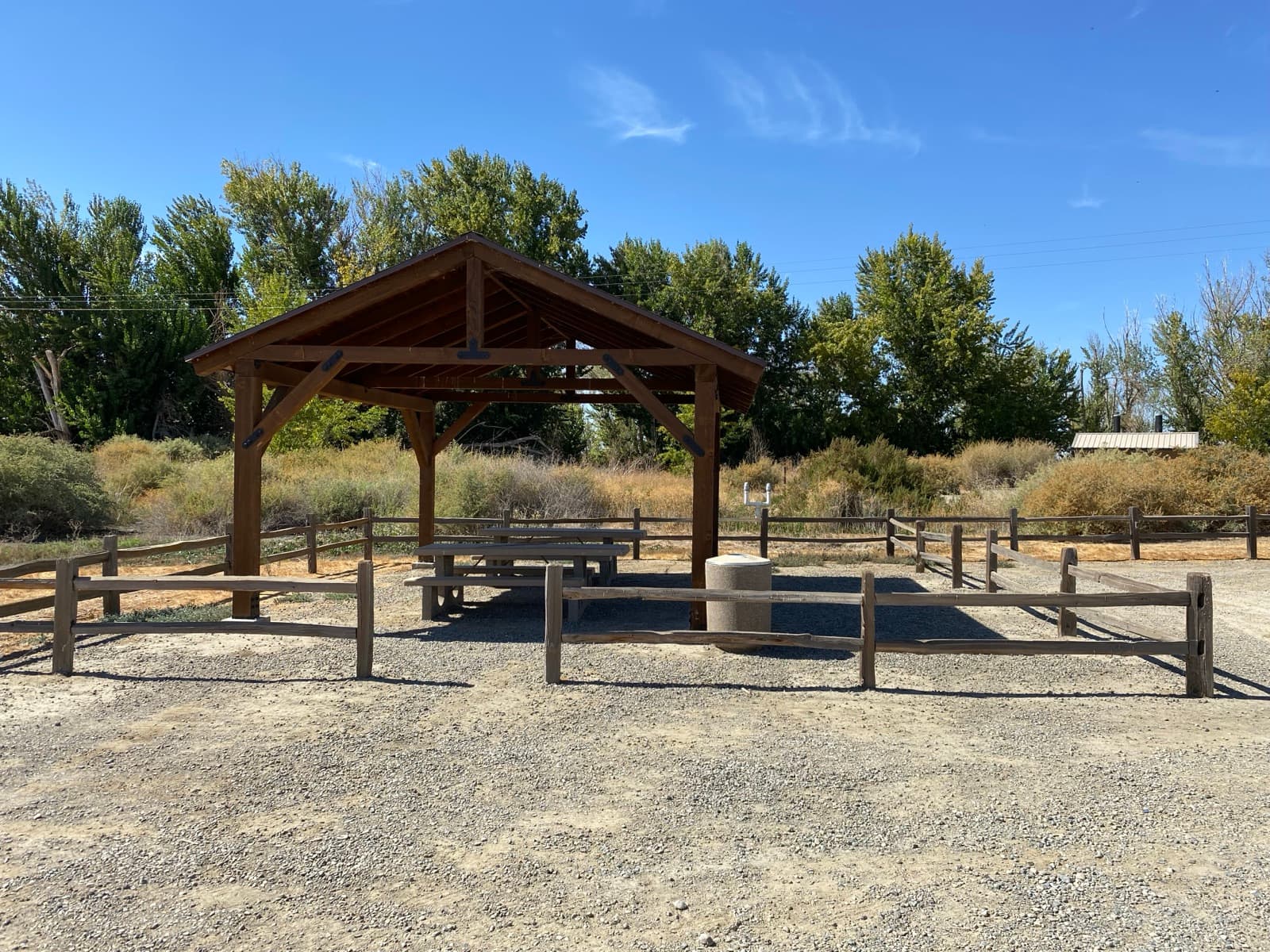 A wooden picnic shelter by a fenced gravel lot, cottonwood trees behind