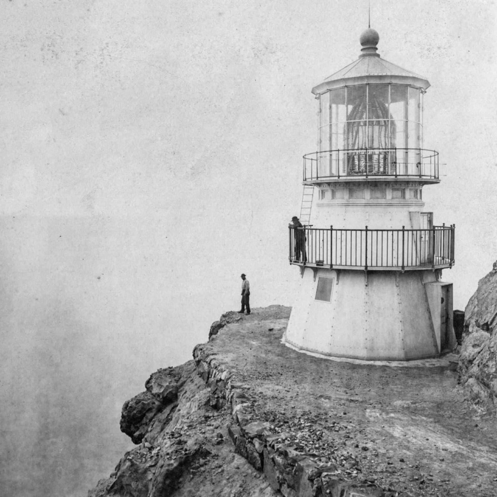 A black and white photo of a short, three-story-tall, white lighthouse on the edge of a cliff, with one man on the tower's balcony and another at the cliff edge