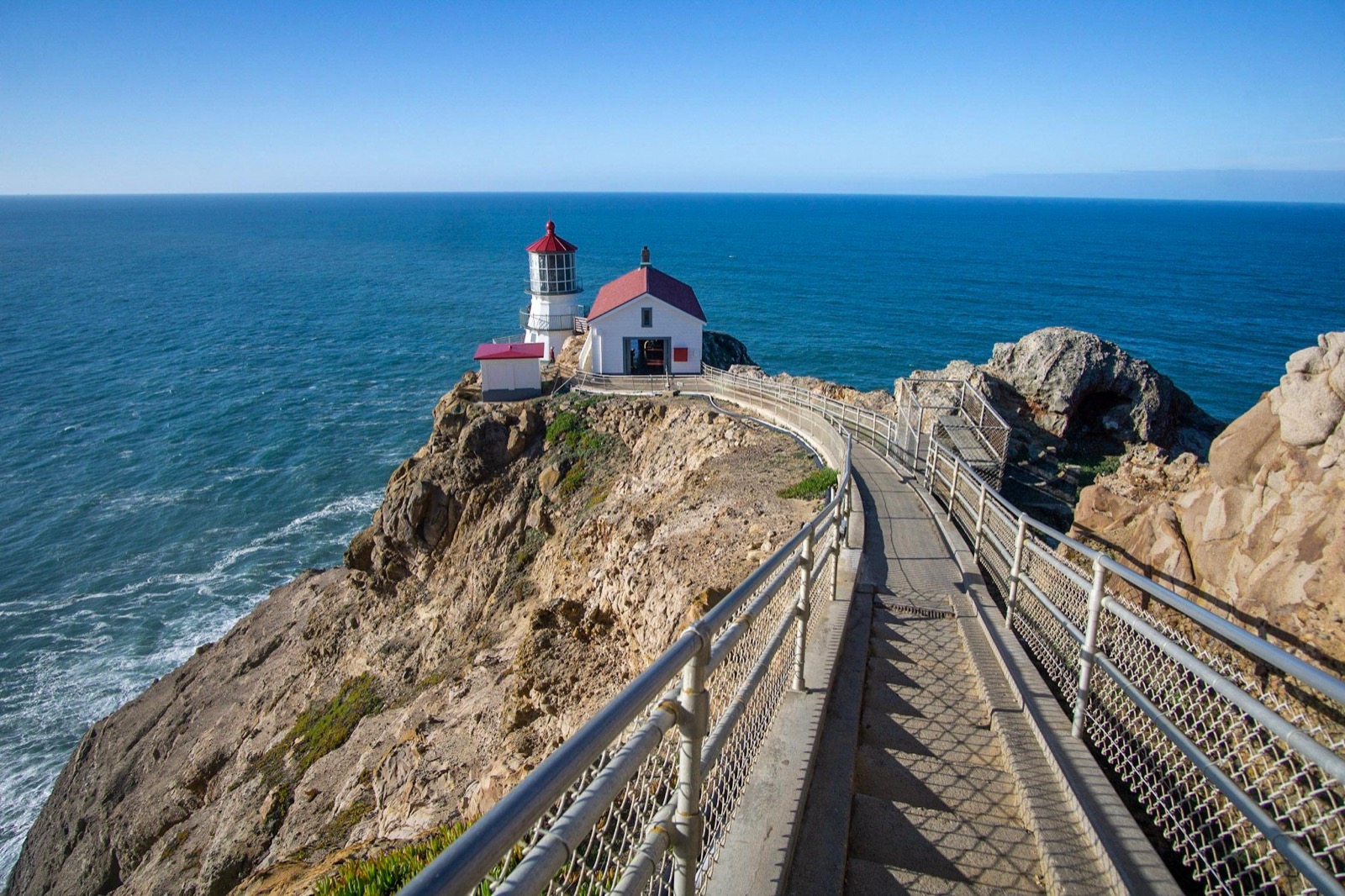 A long concrete staircase leads down to a white lighthouse with a red roof on cliffs above the ocean