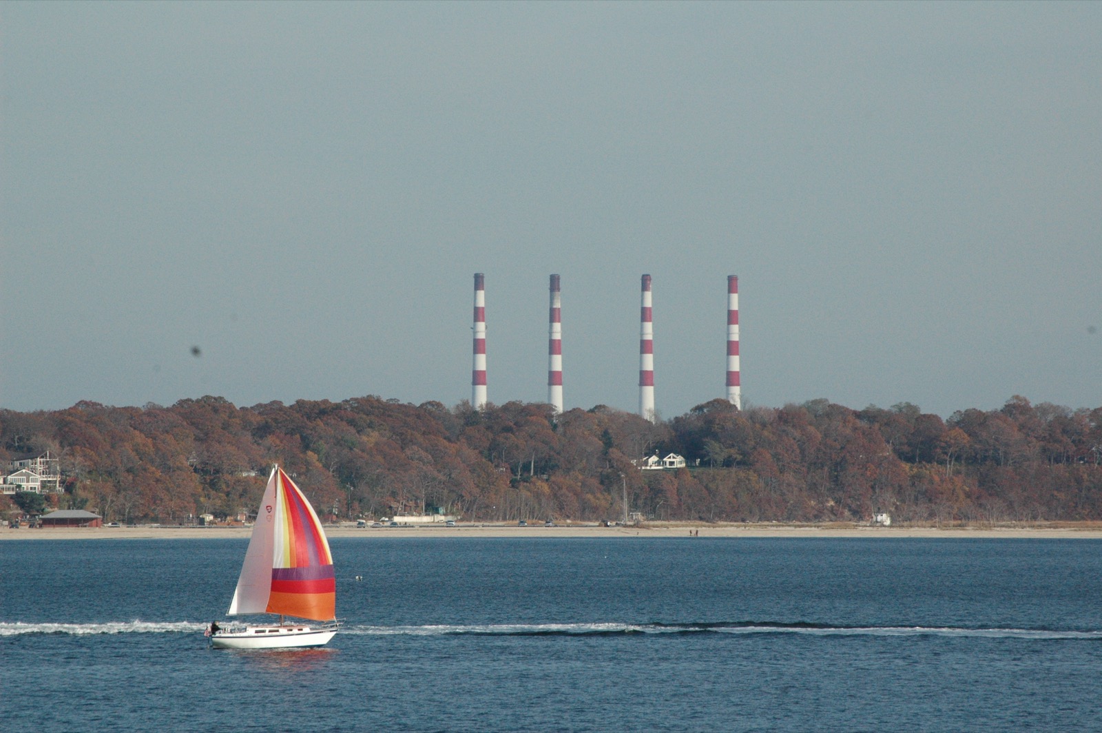 A sailboat with a bright striped sail on blue water, a wooded shoreline and four tall striped smokestacks in the distance