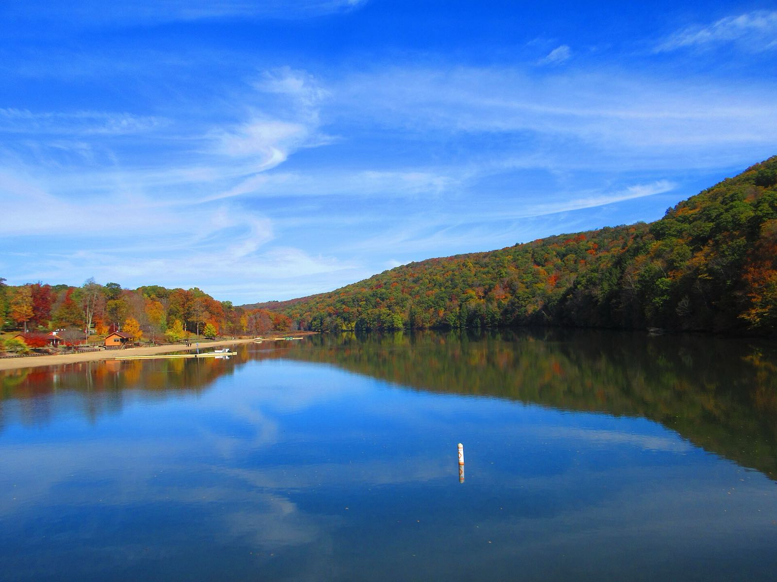 Laurel Hill Lake under a blue sky, with a wooded hillside in mixed red, orange and green fall color reflected in the water and the beach on the left shore