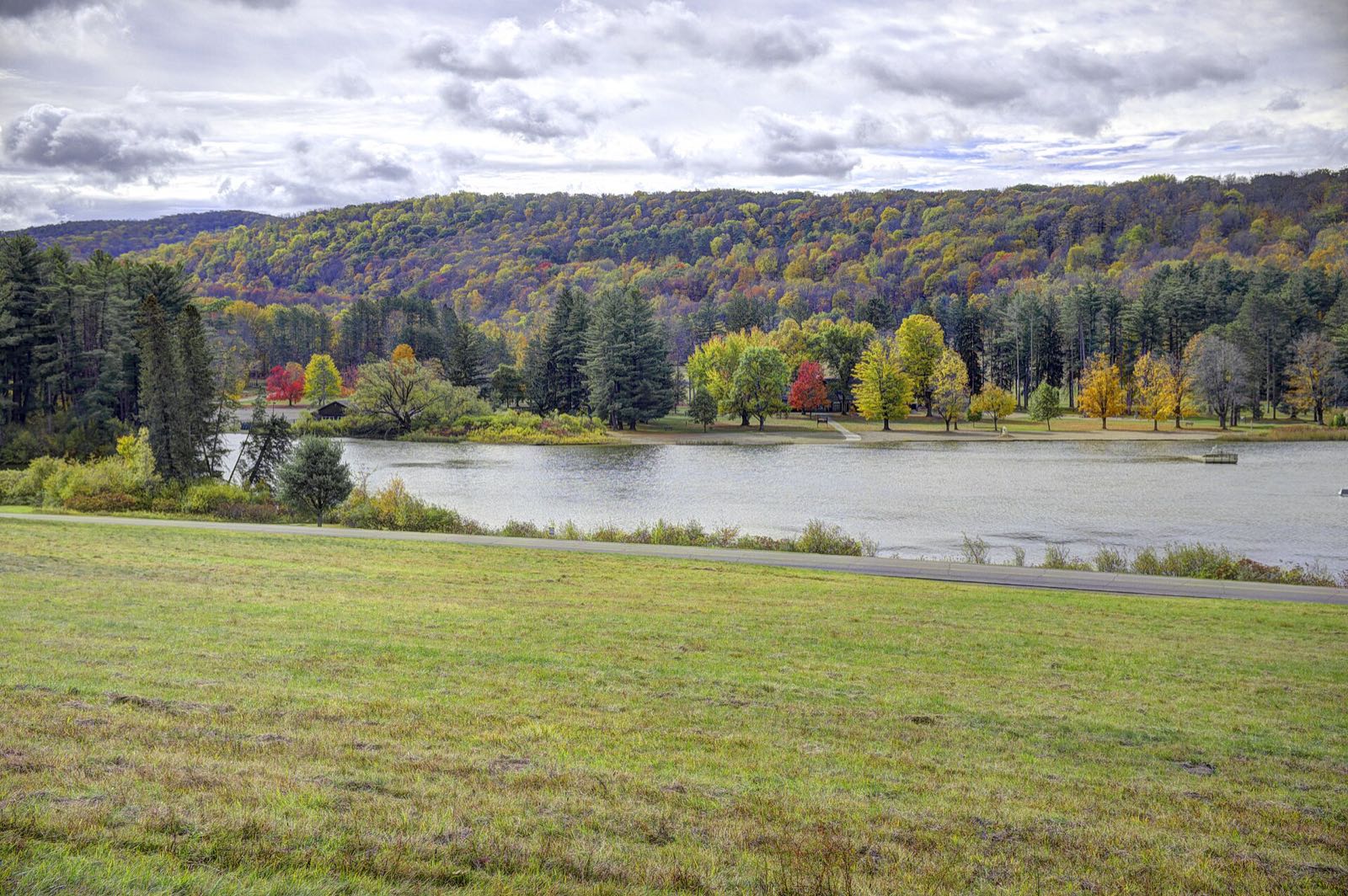 Red House Lake with red, orange and yellow trees along the far shore and a hillside in fall color behind, under a bright cloudy sky; mown grass in front