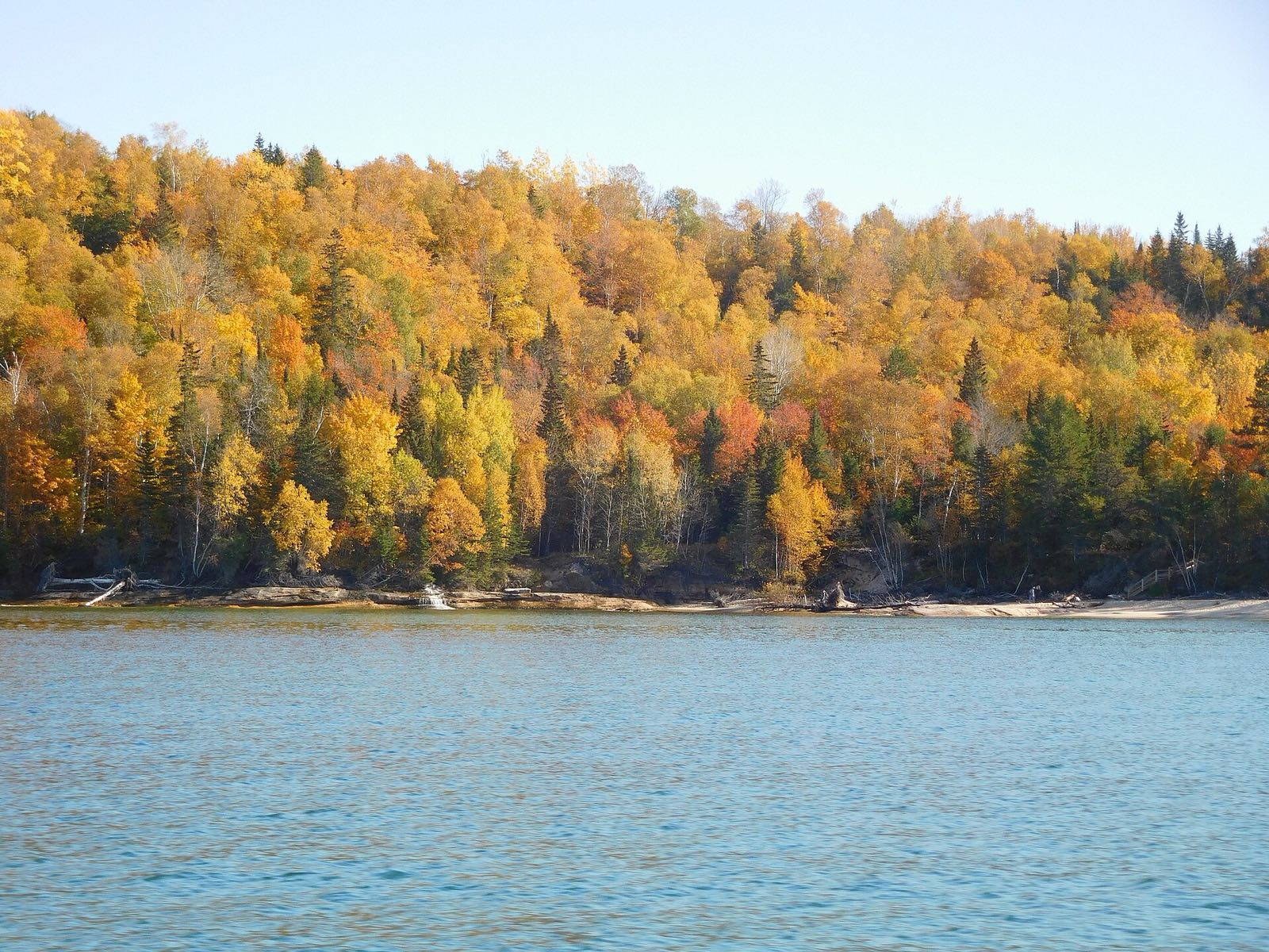 A Lake Superior shoreline backed by a hillside of golden, orange and red birch and maple with dark spruces, calm blue water in front