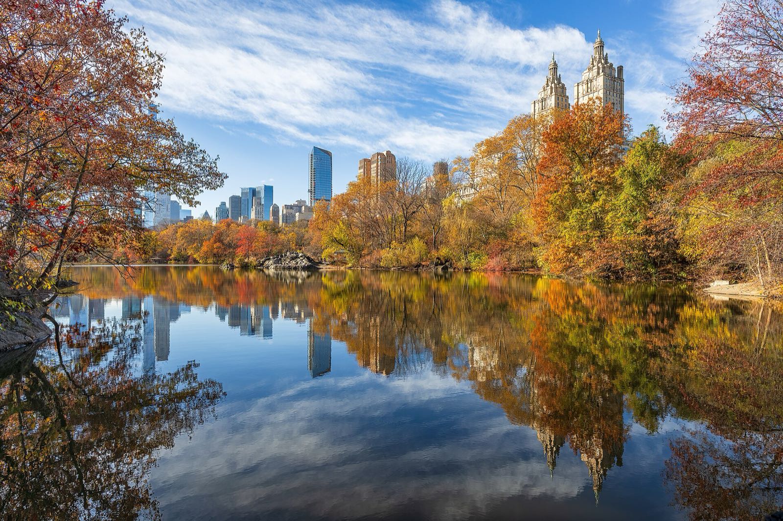 The Lake in Central Park ringed by orange, red and yellow trees, the midtown skyline and the San Remo towers reflected in still water under a blue sky