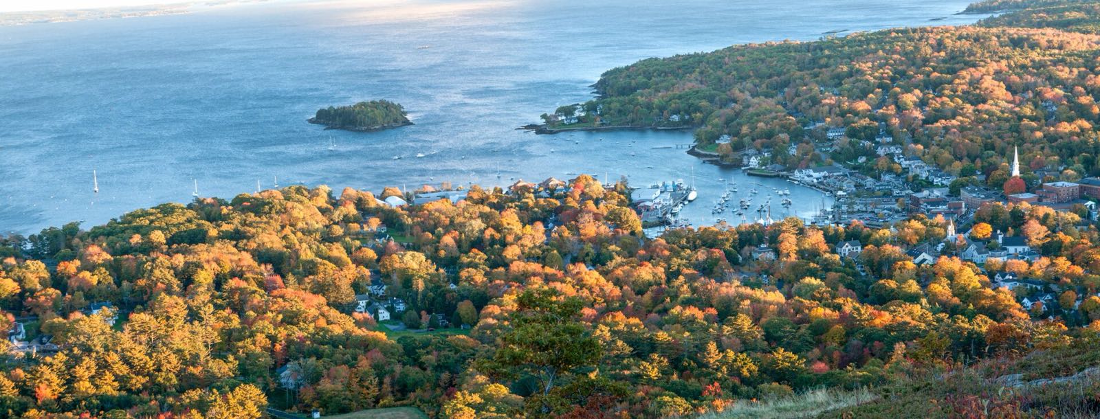 The view from the top of Mount Battie: Camden harbor, the town and its church steeple, and a small island in Penobscot Bay, with trees in gold and orange fall color all around, in late-afternoon light