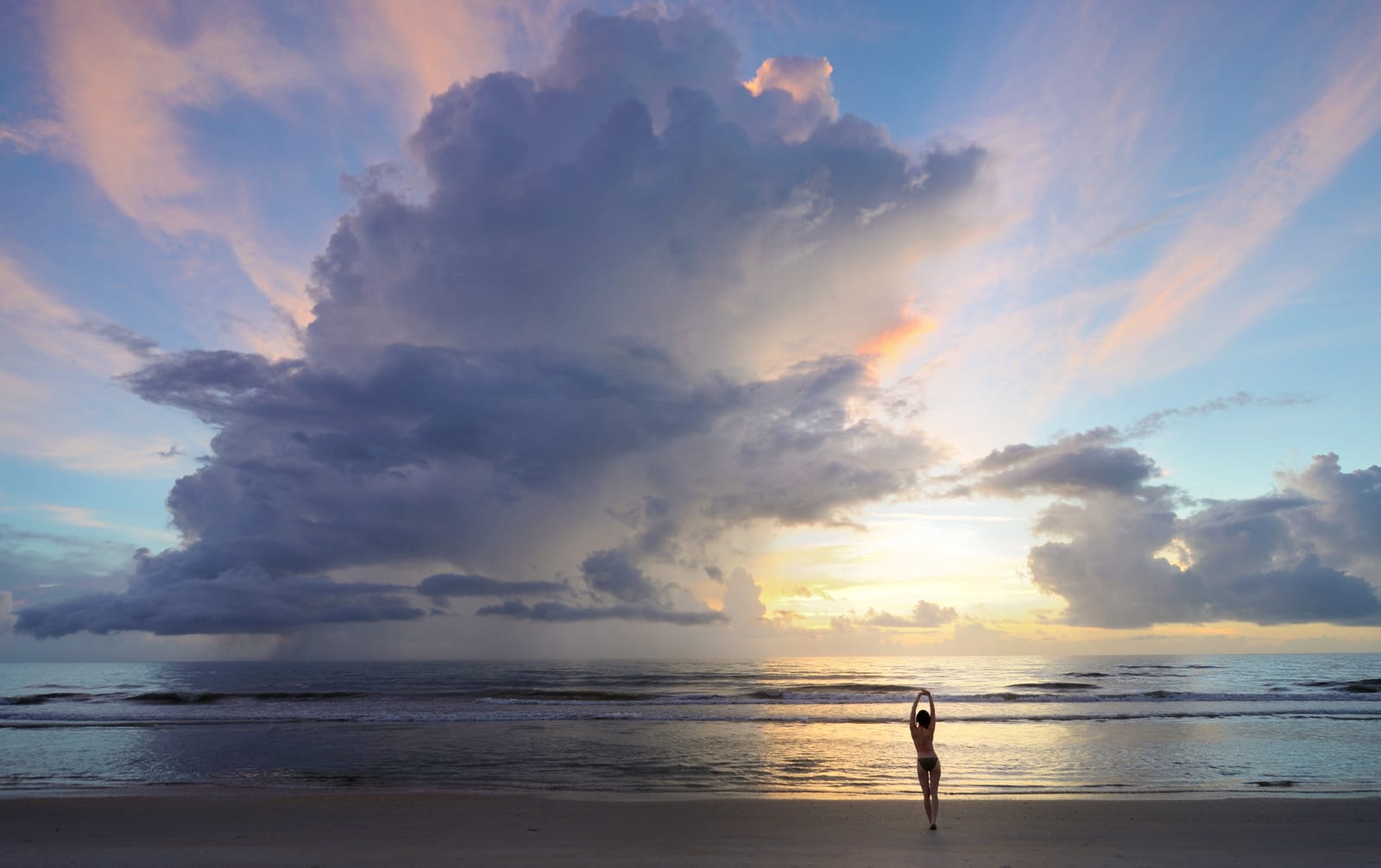 A storm cloud and the rising sun over calm surf, with one person standing at the water's edge