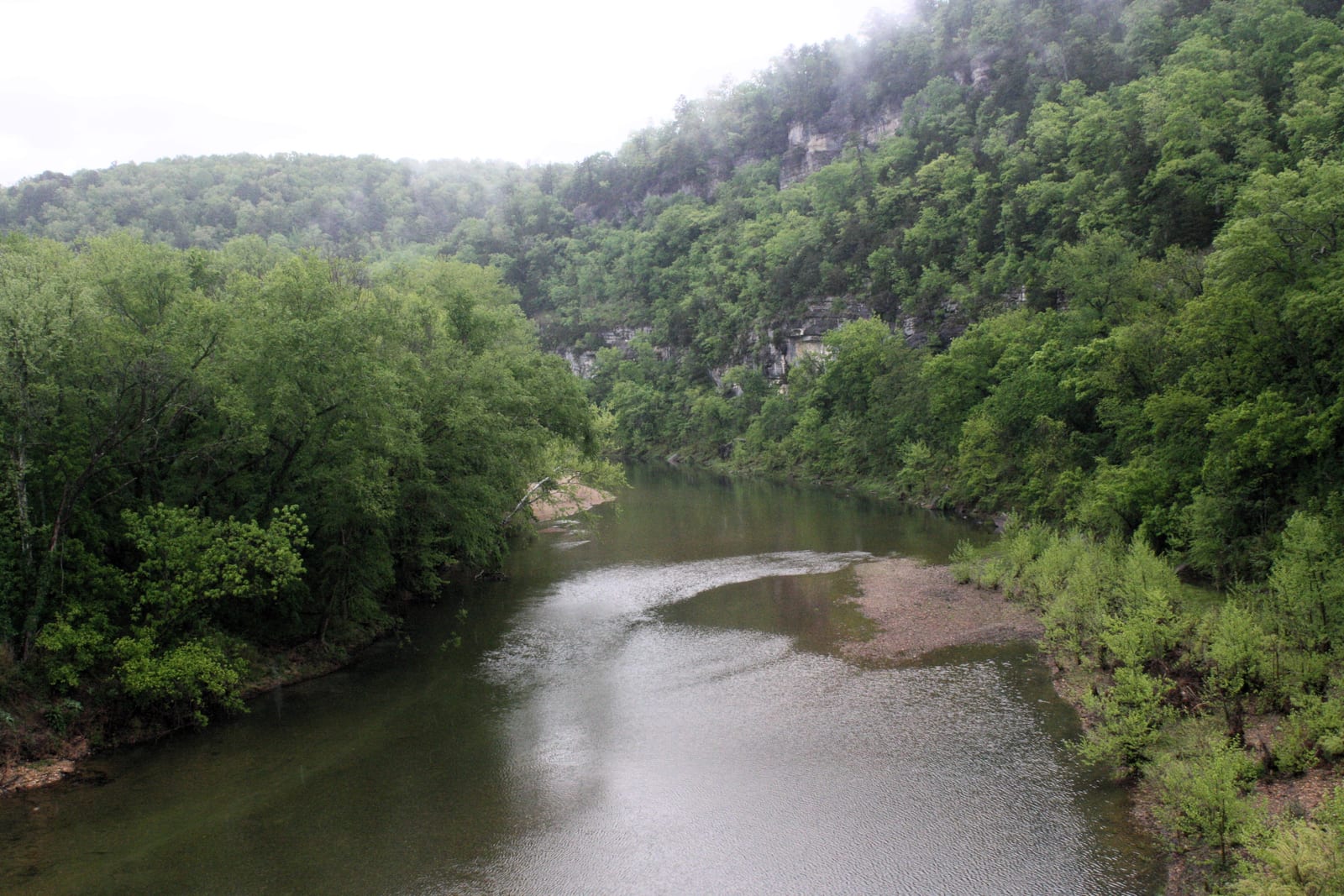 A wide, calm river curving between tree-covered bluffs under a hazy sky